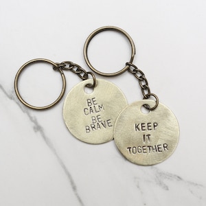 May include: Two gold-toned keychains with round metal tags. One tag says "Be Calm Be Brave" and the other says "Keep It Together".