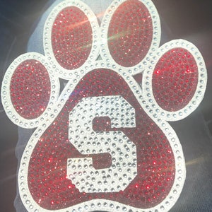 May include: A red paw print with a white letter 'S' in the center. The paw print is covered in small, sparkling rhinestones.