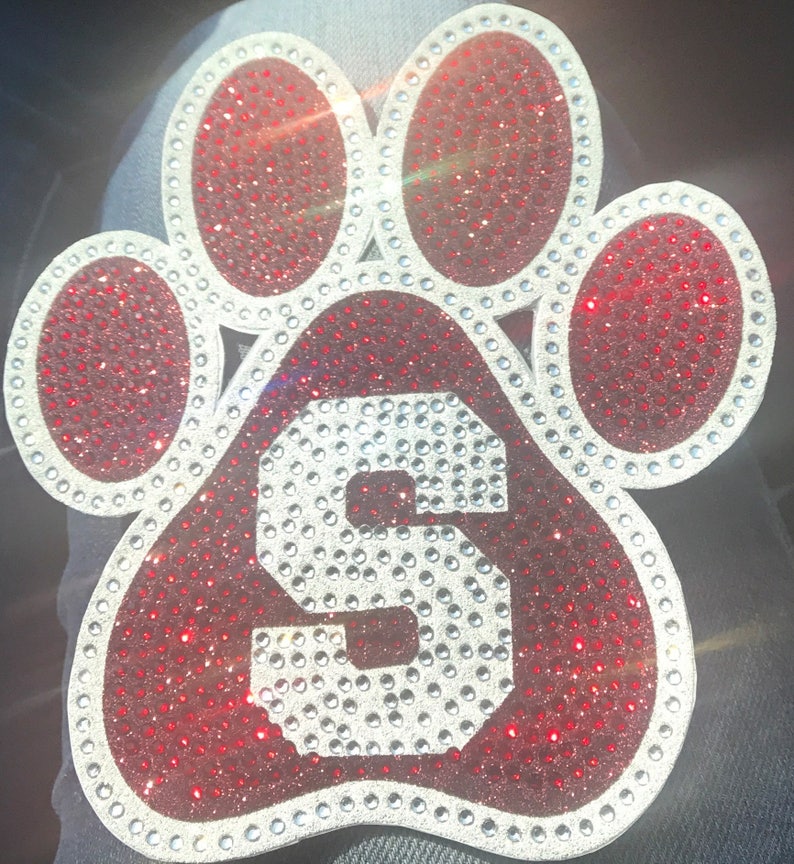 May include: A red paw print with a white letter 'S' in the center. The paw print is covered in small, sparkling rhinestones.
