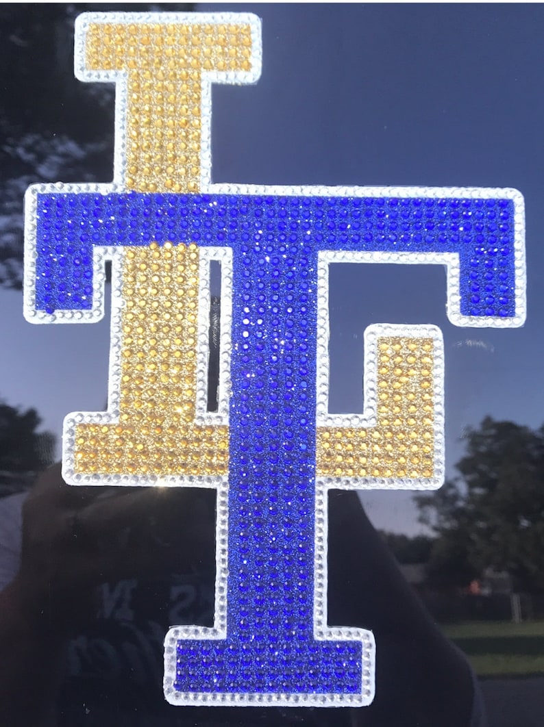 May include: A large, white-outlined, blue and gold letter "T" made of rhinestones. The letter is on a black background.