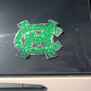 May include: A green and silver car decal featuring the letters "NC" in a stylized font, made of rhinestones.