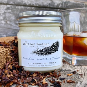 May include: A white soy candle in a glass jar with a silver lid. The label reads "The Ruffled Feather Candle Co." and "Bourbon, Leather, & Tobacco." The candle is surrounded by brown tobacco leaves.