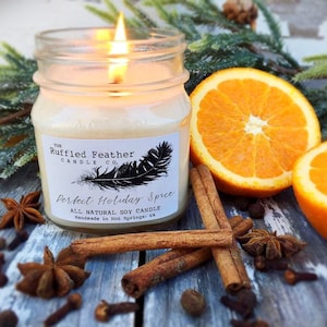 May include: A glass jar candle with a white label that reads "The Ruffled Feather Candle Co. Perfect Holiday Spice All Natural Soy Candle Handmade in Hot Springs, VA". The candle is lit and there are orange slices, cinnamon sticks, and star anise around it.