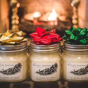 May include: Three glass jars with lids, each topped with a different colored bow. The jars contain soy candles and have labels that read "The Ruffled Feather Candle Co. All Natural Soy Candle Handmade in Hot Springs, AR".
