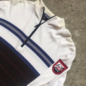 Puede incluir: Camiseta de manga larga a rayas blancas y negras con un detalle de rayas azules y blancas. La camisa tiene un cuello con cremallera y un parche con el texto "Ralph Lauren Polo Sport" y una pequeña bandera estadounidense.