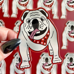 May include: A white bulldog sticker with black detailing, wearing a red bandana with white paw prints. The sticker is held against a red background with multiple bulldog stickers.