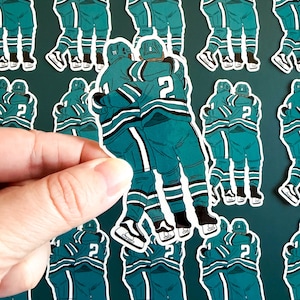 Könnte beinhalten: Eine Hand hält einen Aufkleberbogen mit Illustrationen von Eishockeyspielern in türkis-weißen Uniformen. Die Aufkleber zeigen sich umarmende Spieler, mit der Nummer 2 auf dem Rücken eines Trikots. Die Aufkleber haben einen weißen Rand.