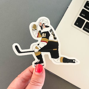 May include: A black and gold hockey player sticker with a white helmet and a stick. The player is on one knee with their arm raised in the air. The player's jersey has the number 81 and the name "Marchessault" on the back.