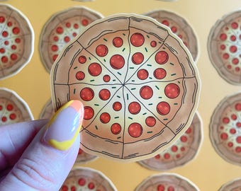 May include: A close-up of a pepperoni pizza sticker with a brown crust and green pepper toppings. The sticker is being held by a hand with colorful nail polish.