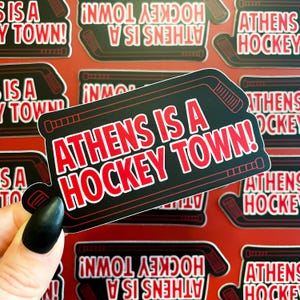 May include: A black and red sticker with the text "Athens is a Hockey Town!" in white. The sticker features a hockey stick graphic.
