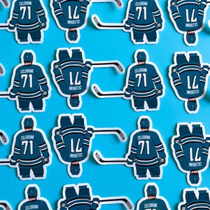 May include: A repeating pattern of teal blue hockey player stickers on a light blue background. Each sticker features a hockey player in a teal blue jersey with the number 71 and the name "CELEBRINI" on the back. The player is in a crouched position with a hockey stick in their hand.