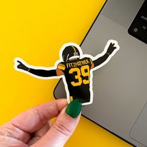May include: A black and yellow football player sticker with the name "Fitzpatrick" and the number "39" on the jersey. The player is in a throwing motion with one arm raised.