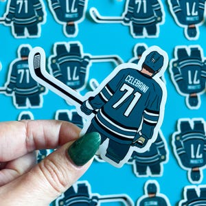 May include: A teal and white hockey player sticker with the name "Celebrini" and the number "71" on the back. The sticker is on a blue background.