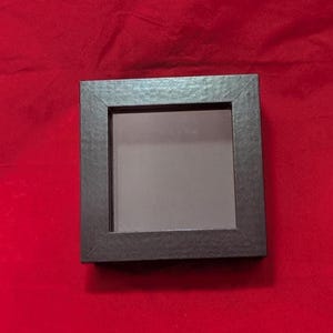 May include: A square, black display box with a textured frame and a smooth, flat center panel. The object features a clean, geometric design with mitered corners on the border, providing a simple, dark, and uniform appearance for the square frame.