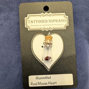 May include: A small clear glass vial pendant with a cork stopper containing a dark red mummified mouse heart. The vial is attached to a black display card featuring the text TATTOOED SOPRANO Honoring our animal friends through art and Mummified Real Mouse Heart.