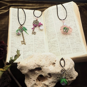 May include: Three handmade pendant necklaces with unique charms, including a key, a flower, and a green and purple design. The necklaces are displayed on an open book and a textured stone, with a dark green background.