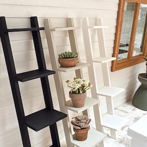 Puede incluir: Tres estanterías de escalera de madera en negro, blanco roto y blanco, cada una con tres estantes. Los estantes están decorados con pequeñas plantas en macetas. El fondo es una pared de madera blanca.