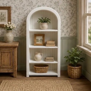 May include: A white arched wooden bookshelf with four shelves featuring vertical beadboard backing. The shelves hold a small potted plant, a framed picture, two stacked wooden boxes, a ceramic pitcher, and a woven basket.