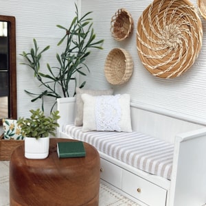 May include: Miniature interior scene with a white daybed featuring a striped cushion and decorative pillows. A round wooden coffee table holds a small green book and a potted plant. Woven wall baskets and a large plant add to the cozy aesthetic.