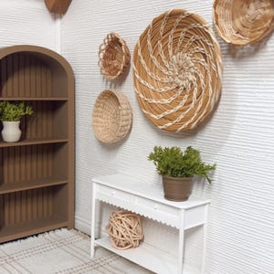May include: A miniature interior scene featuring a white console table, a brown bookshelf, and a woven rug with a plaid pattern. Woven baskets of various sizes are mounted on the textured white wall. A potted plant sits on the table.