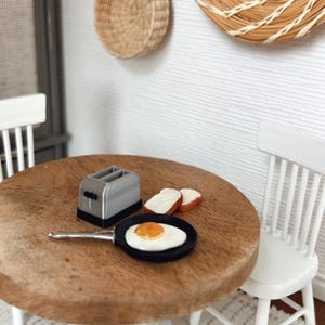 Op de afbeelding: Miniatuur ontbijt met een gebakken ei in een pan, twee sneetjes toast en een zilveren broodrooster op een houten tafel. Twee witte stoelen en wanddecoratie van gevlochten materiaal maken de setting compleet. Een charmante poppenhuiskeuken.