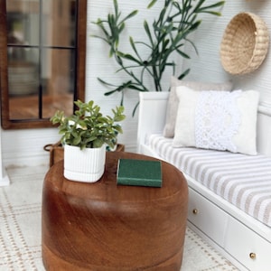May include: Miniature dollhouse interior with a round, dark wood coffee table. A small green book sits on top. A white bench with striped cushions and decorative pillows is in the background. A small potted plant and a window are also visible.