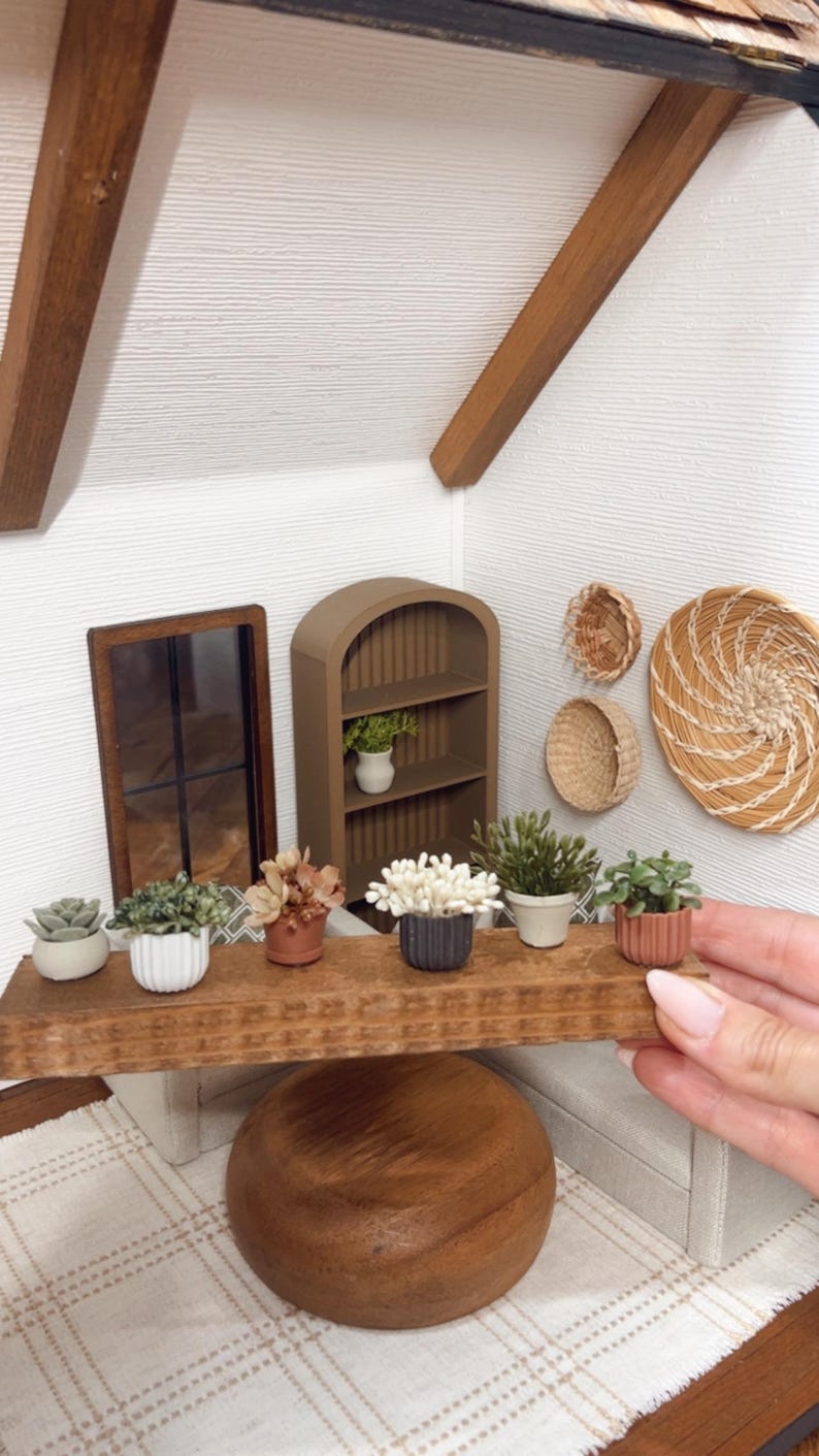 May include: Miniature dollhouse interior with a wooden shelf displaying small potted plants and decorative items. A brown arched bookshelf and a framed mirror are also present. The scene is set against a white textured wall, with wooden beams overhead.
