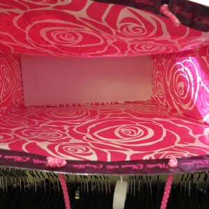 May include: A shopping bag with a vibrant pink interior featuring a white rose pattern. The exterior is dark with pink rope handles. The bag is empty, revealing a white interior. The bag is in a closet.