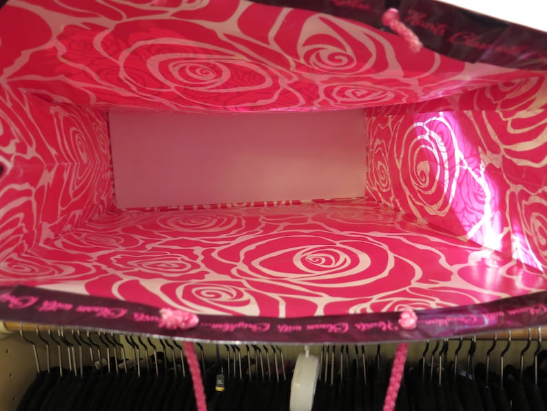 May include: A shopping bag with a vibrant pink interior featuring a white rose pattern. The exterior is dark with pink rope handles. The bag is empty, revealing a white interior. The bag is in a closet.