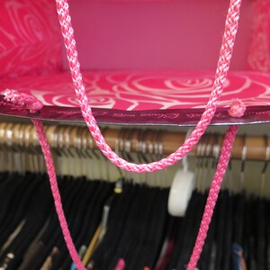 May include: A bright pink gift bag with a braided pink handle. The bag's interior is a solid pink, while the exterior features a white rose pattern. The bag is hanging in a closet with various clothing items.
