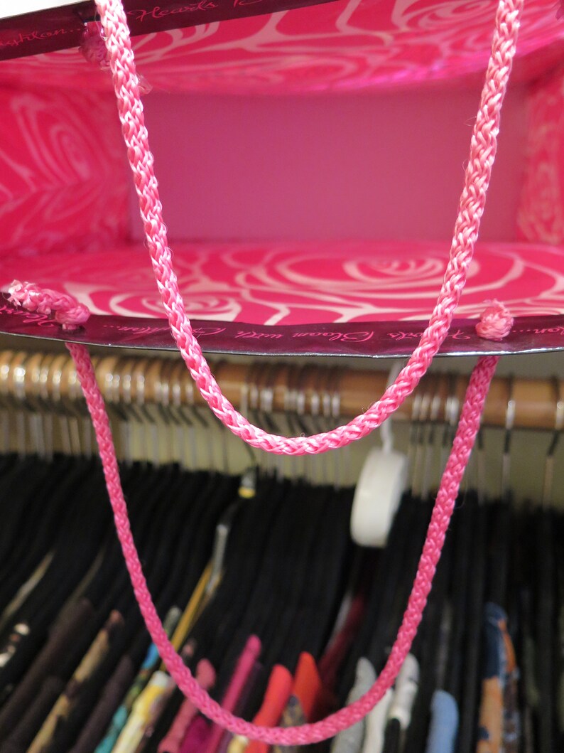 May include: A bright pink gift bag with a braided pink handle. The bag's interior is a solid pink, while the exterior features a white rose pattern. The bag is hanging in a closet with various clothing items.
