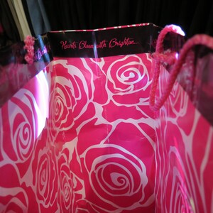 May include: Pink shopping bag with a white rose pattern. The bag has a black trim with the text "Hearts Bloom with Brighton..." and pink rope handles.