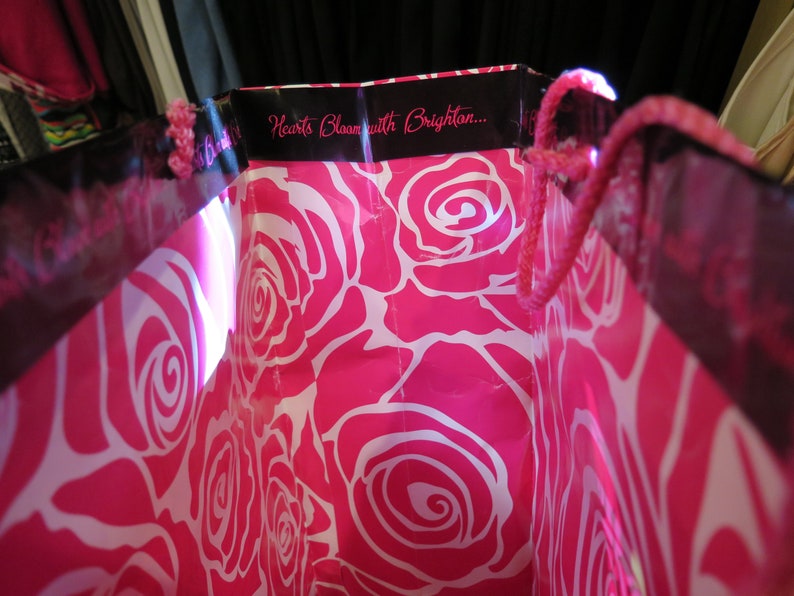 May include: Pink shopping bag with a white rose pattern. The bag has a black trim with the text "Hearts Bloom with Brighton..." and pink rope handles.