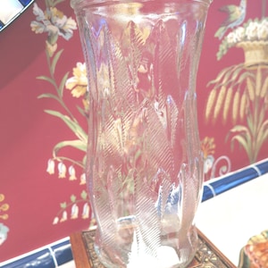 May include: A clear glass vase with a textured, leaf-like pattern. The vase is sitting on a decorative, wooden stand with gold accents.