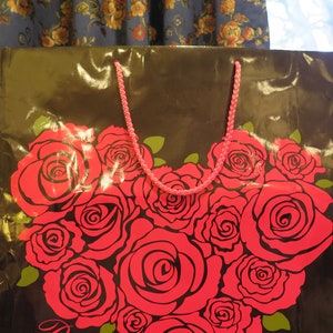 May include: Black gift bag with a heart-shaped arrangement of red roses. The bag has a pink rope handle and green leaves accenting the roses. The bag is shiny and has a floral design.