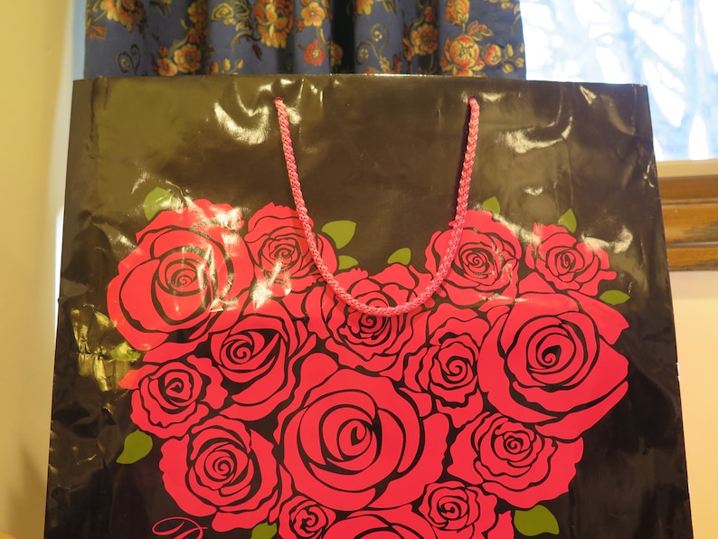 May include: Black gift bag with a heart-shaped arrangement of red roses. The bag has a pink rope handle and green leaves accenting the roses. The bag is shiny and has a floral design.