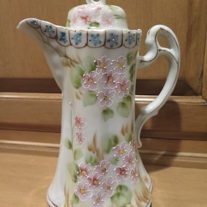 Puede incluir: Una tetera de porcelana blanca con un diseño floral. La tetera tiene una tapa con un diseño floral y un asa. La tetera está decorada con flores rosas y verdes.
