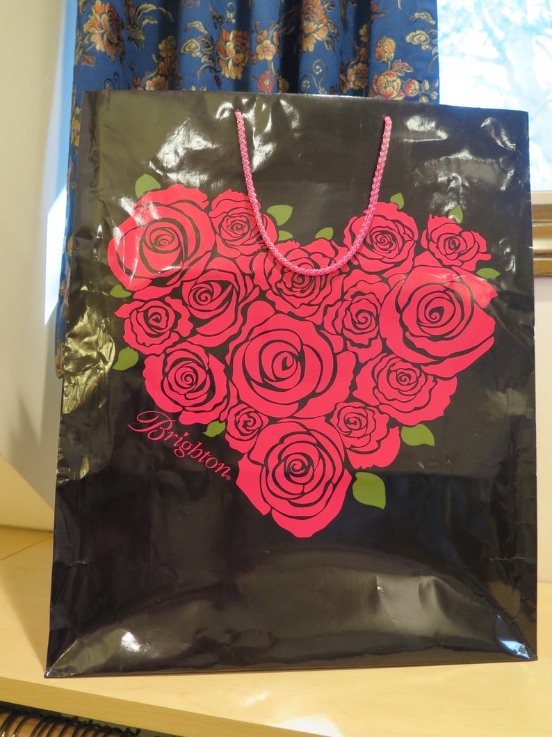 May include: Black gift bag with a heart-shaped arrangement of red roses. The bag has a pink rope handle and the word "Brighton" printed in cursive. The roses have green leaves.