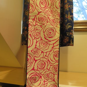 May include: Tall gift bag with a white background and a repeating pattern of pink roses. The bag is rectangular and stands upright, with a black border. The bag is made of paper and is likely used for presenting gifts.