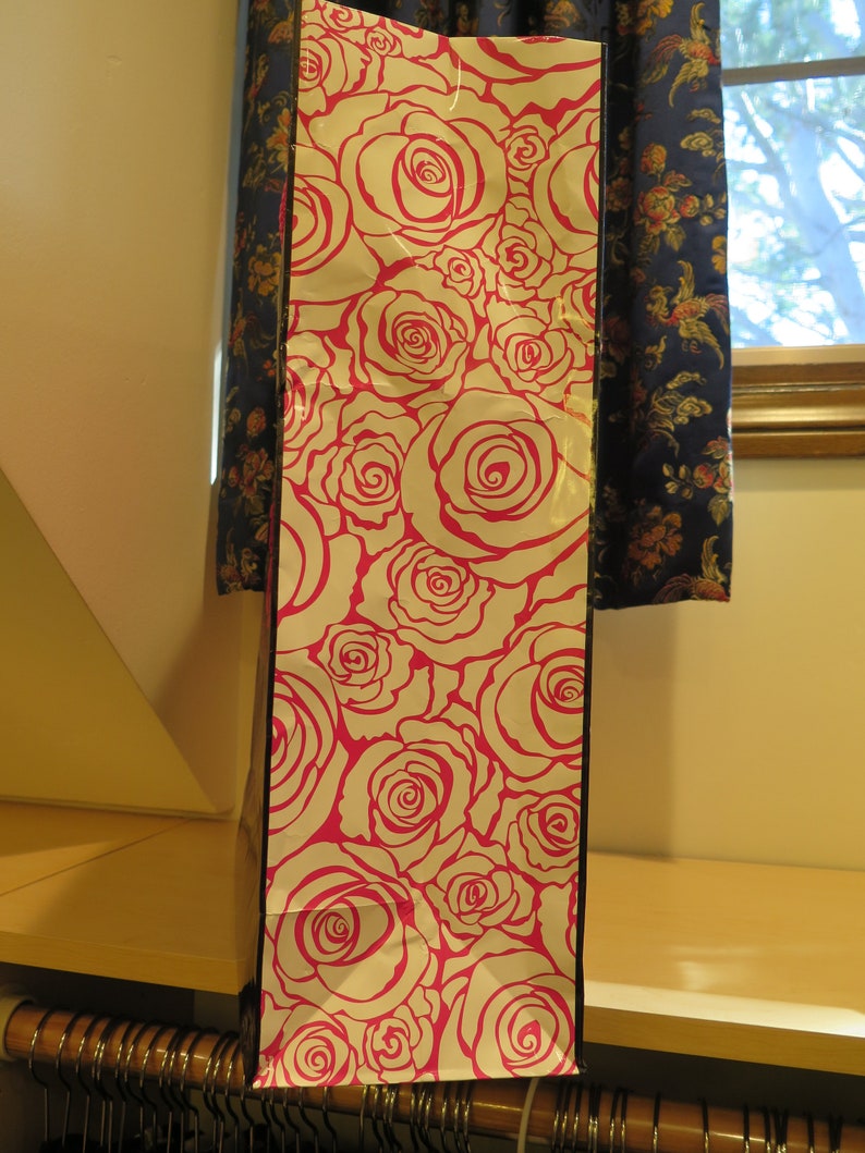 May include: Tall gift bag with a white background and a repeating pattern of pink roses. The bag is rectangular and stands upright, with a black border. The bag is made of paper and is likely used for presenting gifts.