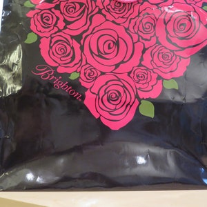 May include: Black shopping bag with a heart-shaped design of pink roses and green leaves. The word "Brighton" is printed in cursive on the bag. The bag has a shiny, reflective surface.