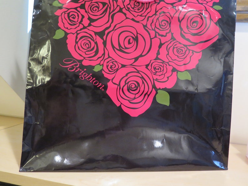 May include: Black shopping bag with a heart-shaped design of pink roses and green leaves. The word "Brighton" is printed in cursive on the bag. The bag has a shiny, reflective surface.