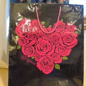 May include: Black shopping bag with a heart-shaped design of pink roses and green leaves. The bag has a pink rope handle and the word "Brighton" in cursive. The bag is made of a shiny material.