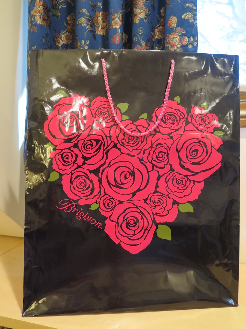 May include: Black shopping bag with a heart-shaped design of pink roses and green leaves. The bag has a pink rope handle and the word "Brighton" in cursive. The bag is made of a shiny material.