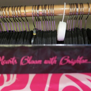 May include: A closet filled with black clothing hanging on metal hangers with purple accents. The hangers are suspended from a wooden rod. A sign with the text "Hearts Bloom with Brighton..." is visible.