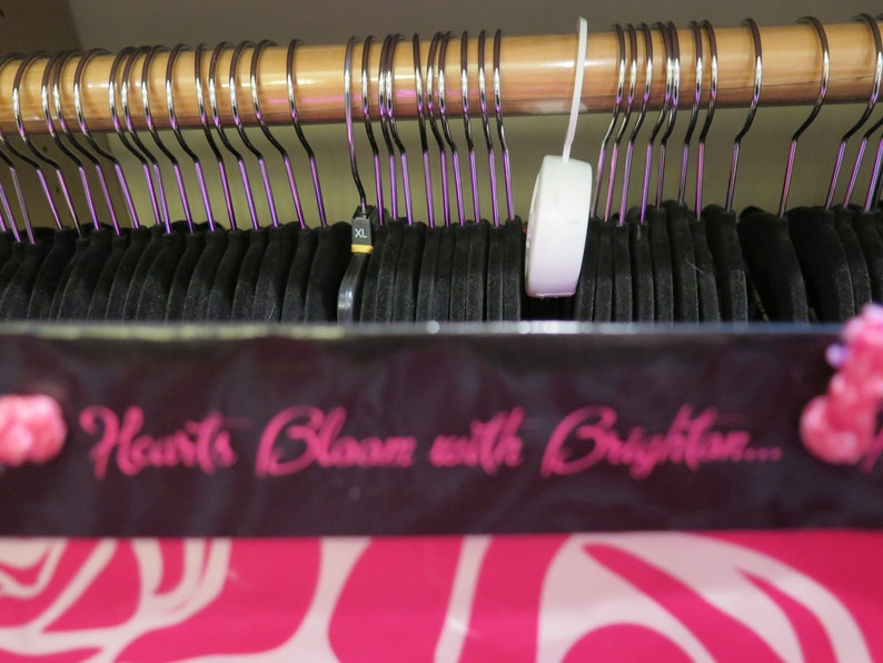 May include: A closet filled with black clothing hanging on metal hangers with purple accents. The hangers are suspended from a wooden rod. A sign with the text "Hearts Bloom with Brighton..." is visible.