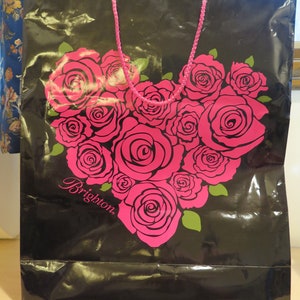May include: Black shopping bag with a heart-shaped arrangement of pink roses. The bag has a pink rope handle and the word "Brighton" in cursive. Green leaves accent the roses.