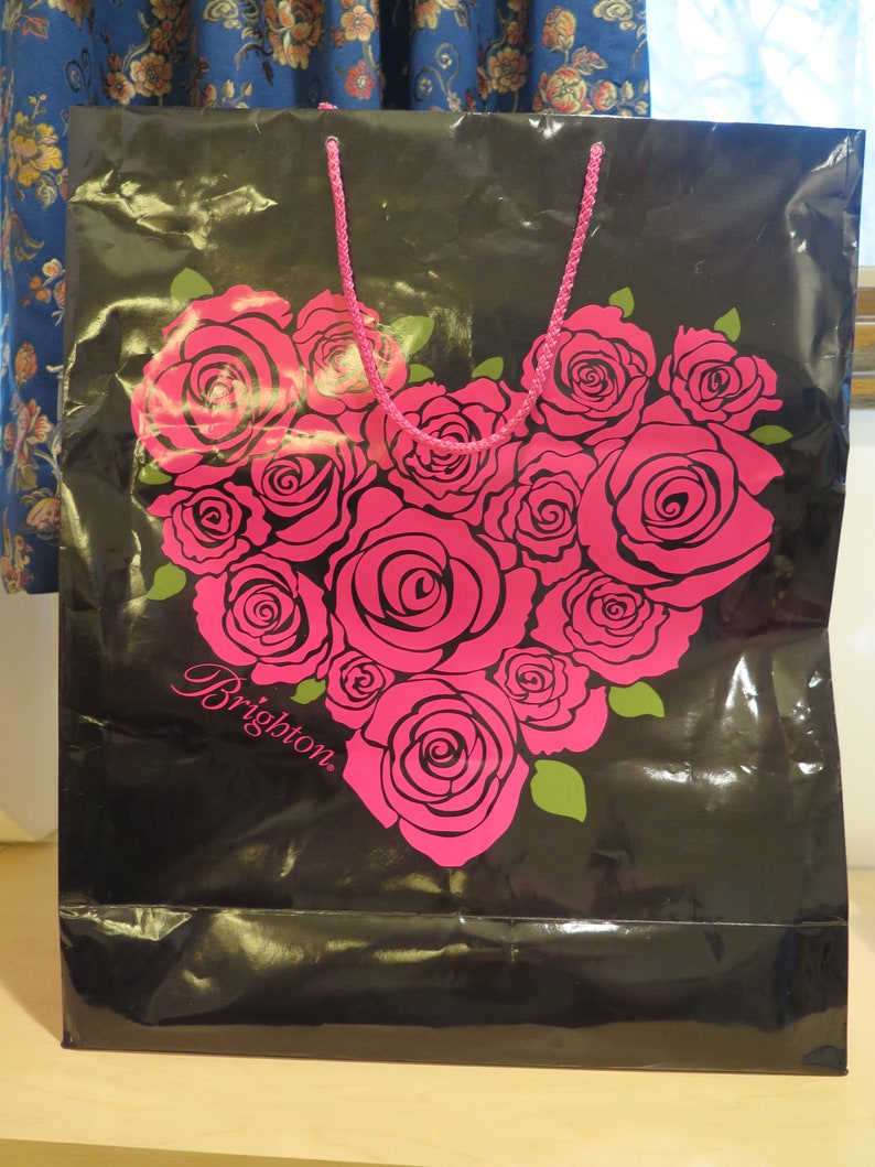 May include: Black shopping bag with a heart-shaped arrangement of pink roses. The bag has a pink rope handle and the word "Brighton" in cursive. Green leaves accent the roses.