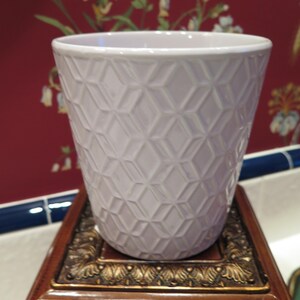 Puede incluir: Una maceta de cerámica lila claro con un patrón de diamantes geométricos. Se apoya en un soporte de madera decorativo con adornos dorados. El fondo presenta un papel pintado floral.