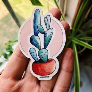 Puede incluir: Una pegatina con una ilustraci&oacute;n de acuarela de un cactus azul en una maceta roja, sobre un fondo rosa. La pegatina tiene un borde blanco y el texto "oneloverartshop" en la parte inferior.